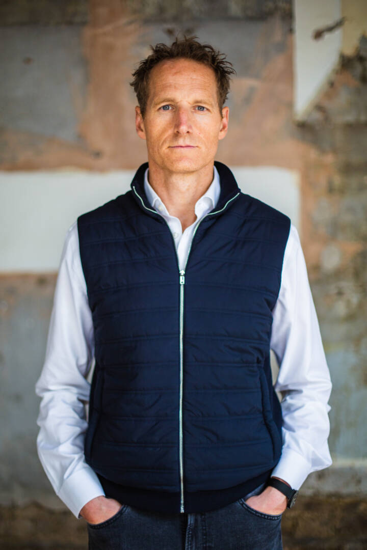 Lifestyle portrait off man in blue puffer gillet with hands in jeans pockets against an exposed brick wall by portrait photographer Andy Barnham