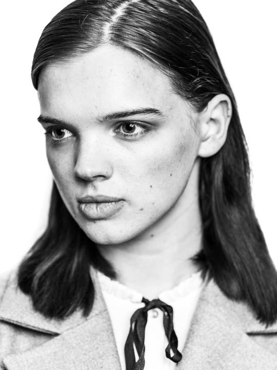 Portrait of a woman with long hair in overcoat and white shirt with a bow in front of a white background by portrait photographer Andy Barnham