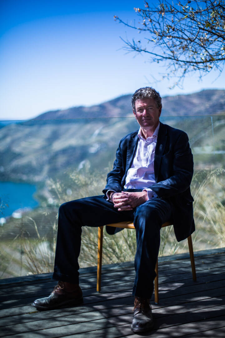 Lifestyle portrait of managing director of Quinta do Noval Christian Seely hands in his lap, sitting on on a chair on a deck with sunny river and valley in background by portrait photographer Andy Barnham