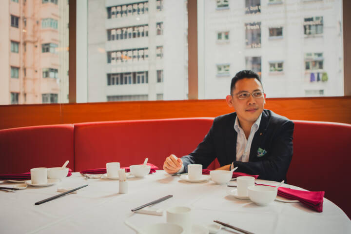 Lifestyle portrait of co-founder of the World of Mouth food guide Fergus Fung with glasses sitting at an orange coloured booth at a restaurant table laid out with bowls and chopsticks with residential high rise buildings in the background by portrait photographer Andy Barnham