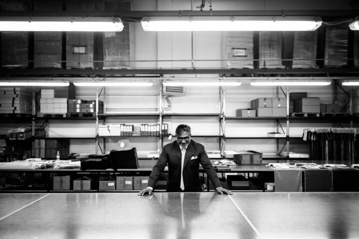 Lifestyle portrait of man with glasses in suit leaning on large table with both hands in a warehouse by portrait photographer Andy Barnham