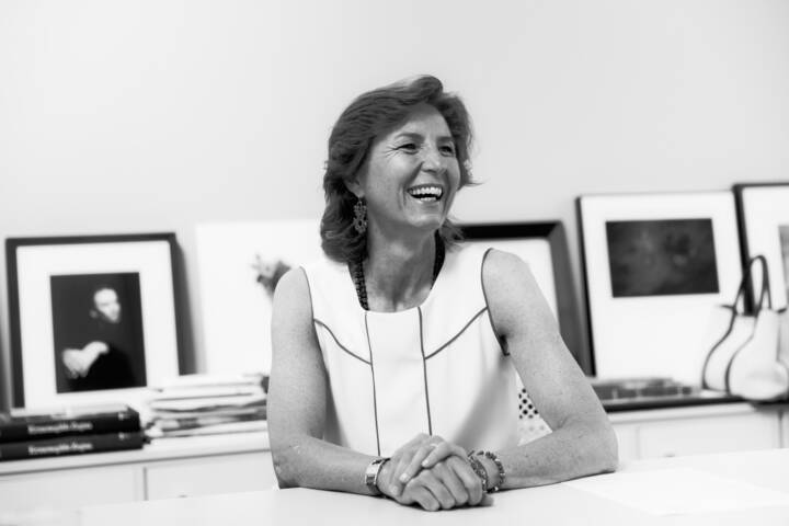 Lifestyle portrait of president of Fondazione Zegna Anna Zegna laughing in a white dress sitting at a table with framed photos on shelf behind by portrait photographer Andy Barnham