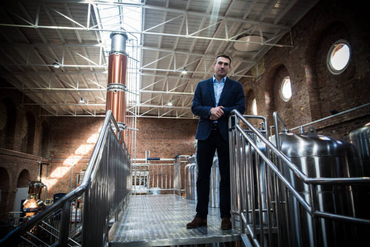 Lifestyle portrait of co-founder of the Copper Rivet Distillery Stephen Russell in jacket and shirt standing in front of a copper still and whisky mash in a distillery by portrait photographer Andy Barnham