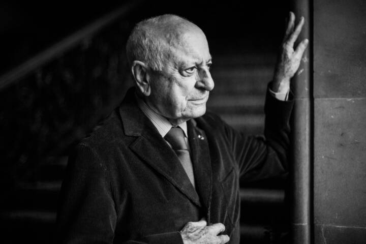 Lifestyle portrait of co-founder fashion label Yves Saint Laurent Pierre Berge in jacket shirt and tie looking out a window with hand on the window frame with staircase behind by portrait photographer Andy Barnham