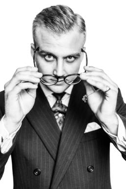 Portrait expressions; Joe Holsgrove, portrait of the the Savile Row tailor wearing a suit and looking at the camera while putting his glasses on