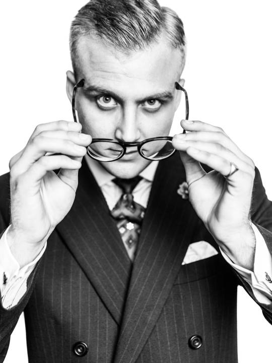 Portrait of a man in a suit a tie putting on a pair of glasses in front of a white background by portrait photographer Andy Barnham