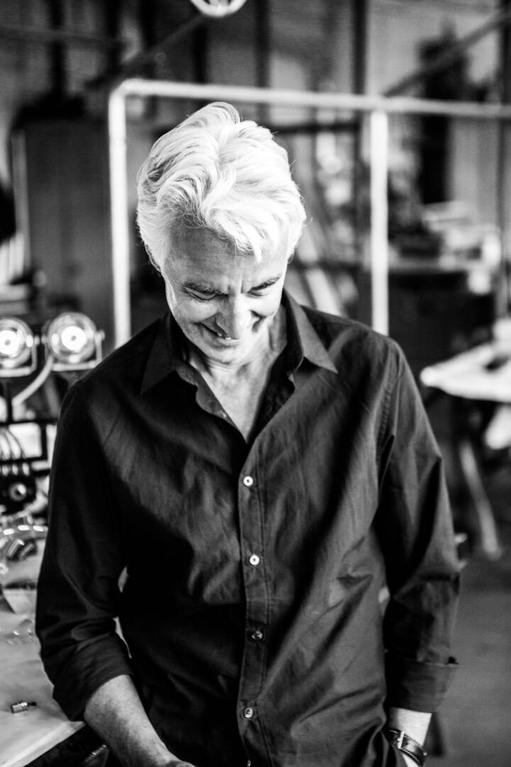 Lifestyle portrait of industrial designer Frank Buchwald smiling in a black shirt looking at the floor in an industrial workshop by portrait photographer Andy Barnham