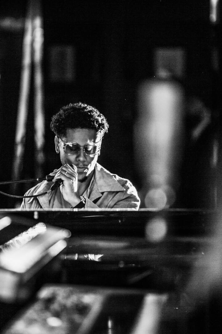 Lifestyle portrait of British singer and songwriter Reuben James in glasses sitting at piano holding microphone by portrait photographer Andy Barnham