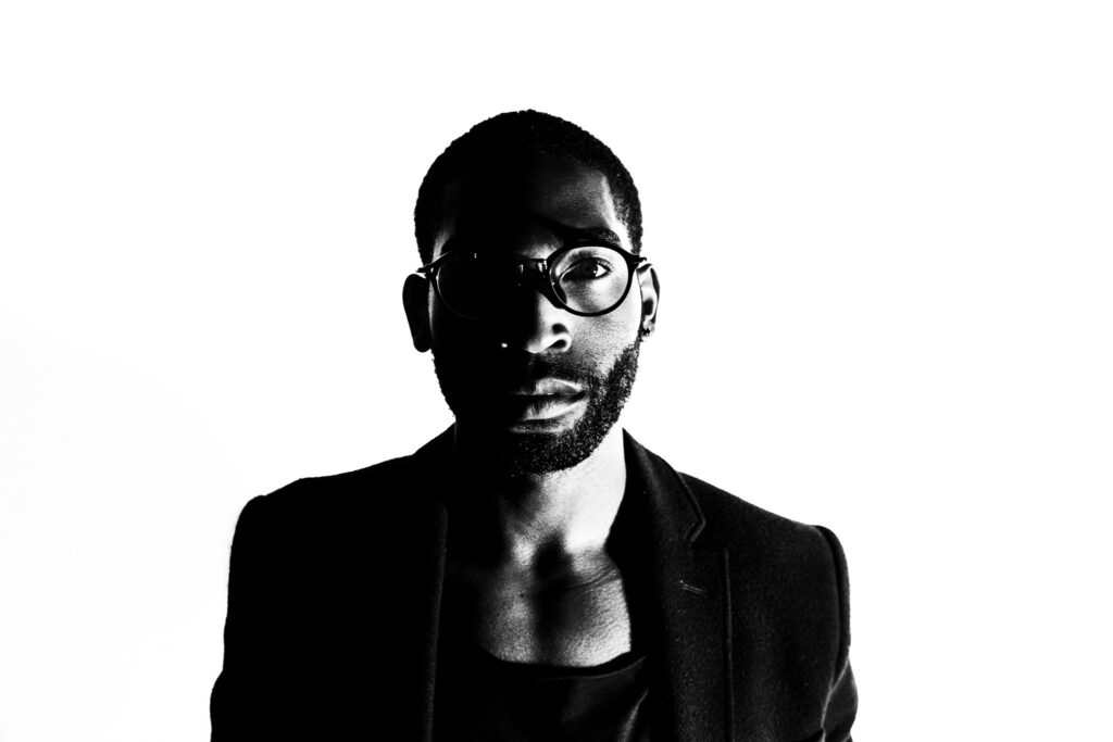 Black and white studio portrait of British rapper and singer Tinie Tempah in glasses and a jacket looking at the camera lit by one light on camera right by portrait photographer Andy Barnham