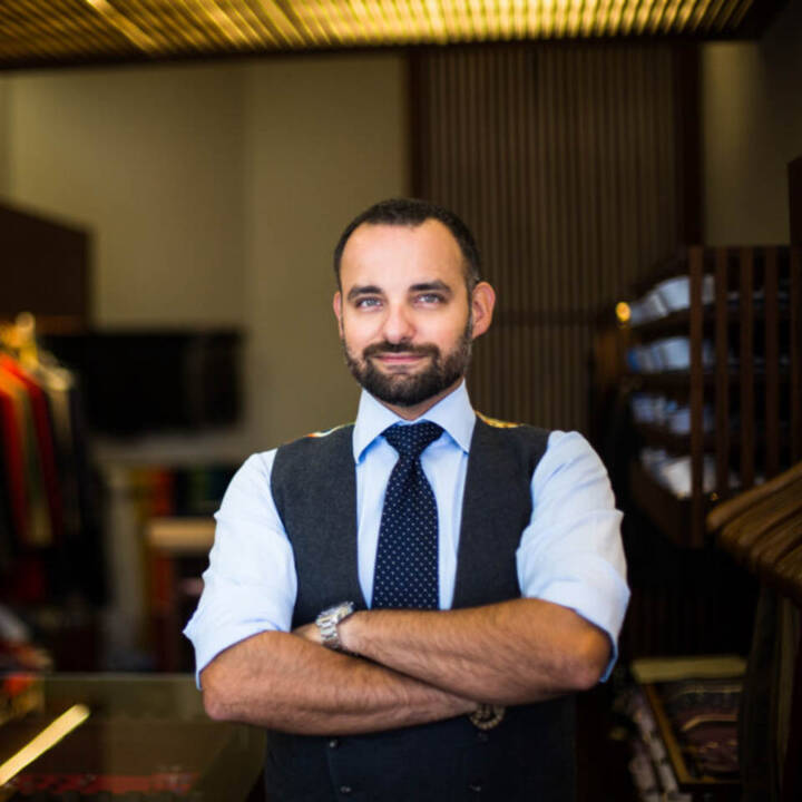Lifestyle portrait of bearded man with arms folded in pale blue shirt dark blue tie and waistcoat standing in menswear store by portrait photographer Andy Barnham