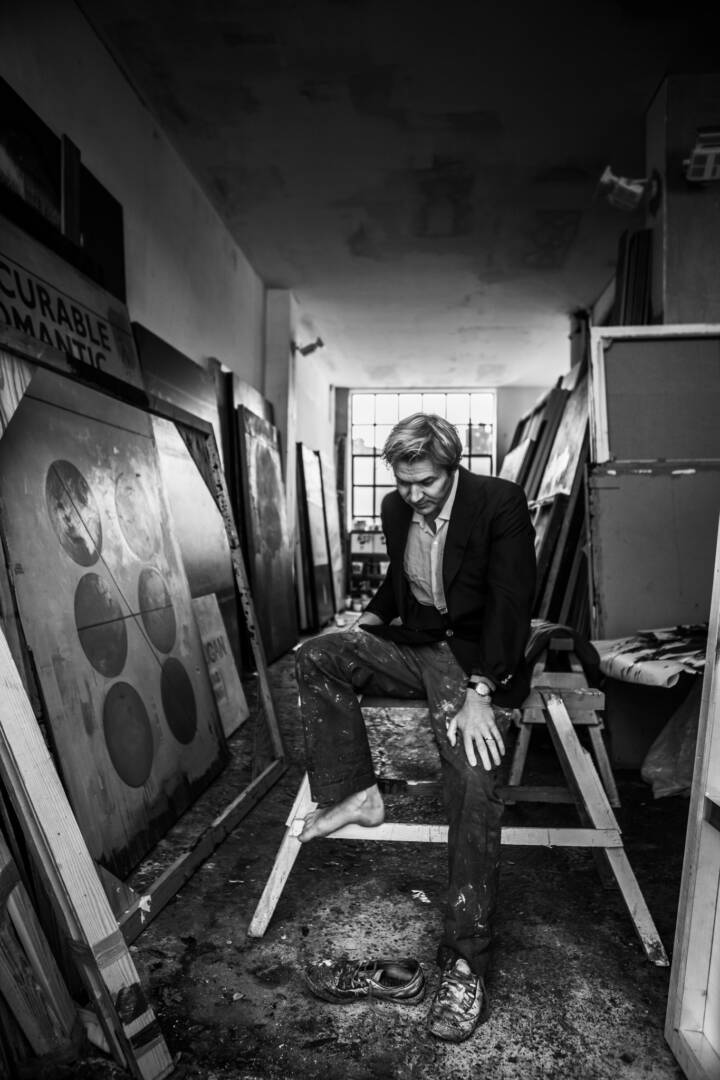 Lifestyle portrait of British writer and artist Harland Miller sitting on a bench in narrow studio lined by paintings by portrait photographer Andy Barnham