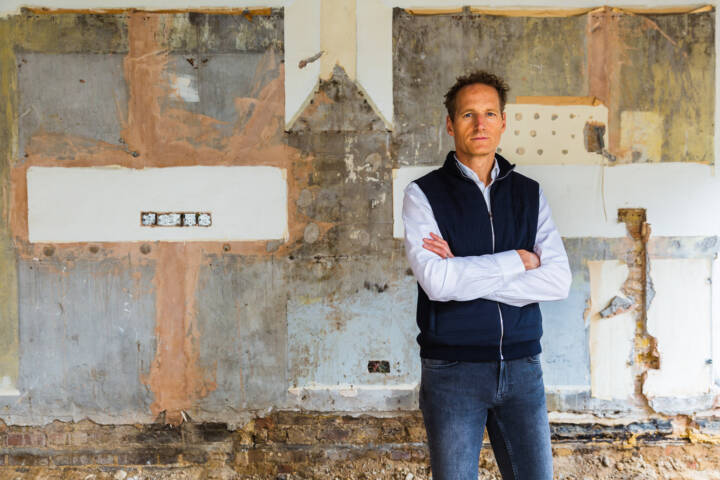 Lifestyle portrait off man in blue puffer gillet with arms folder against an exposed wall by portrait photographer Andy Barnham