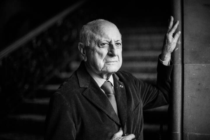 Lifestyle portrait of co-founder fashion label Yves Saint Laurent Pierre Berge in jacket shirt and tie looking out a window with hand on the window frame with staircase behind by portrait photographer Andy Barnham