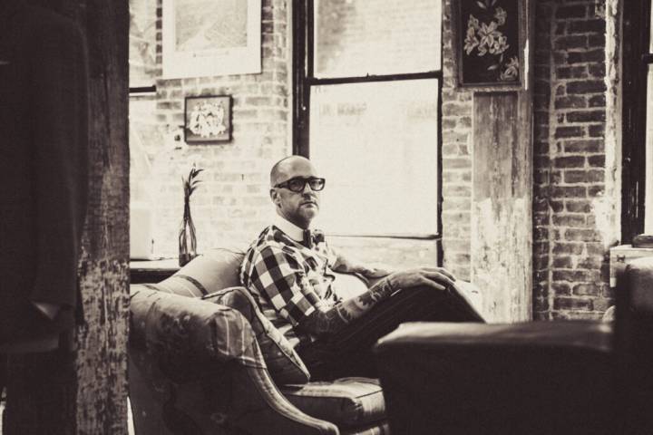 Lifestyle portrait of designer and owner of bespoke tailor Alex Sumner with bow tie in glasses and check shirt showing arm tattoos sitting on a sofa in wood and brick atelier by portrait photographer Andy Barnham