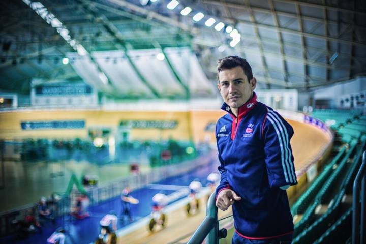 Lifestyle portrait of three-time Paralympic medalist Jaco van Gass with one arm in cycling velodrome with cyclists racing in background by portrait photographer Andy Barnham