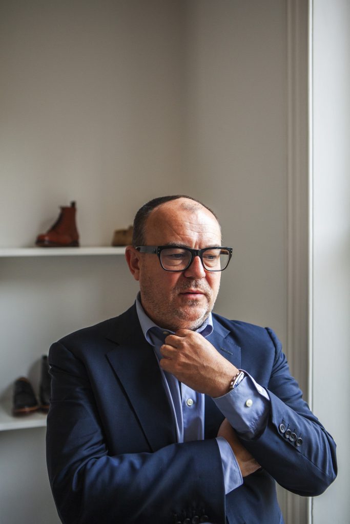 Lifestyle portrait of Tim Little, Creative Director and owner of Grenson and Tim Little with glasses, jacket and shirt standing next to a window playing with his top shirt button by portrait photographer Andy Barnham