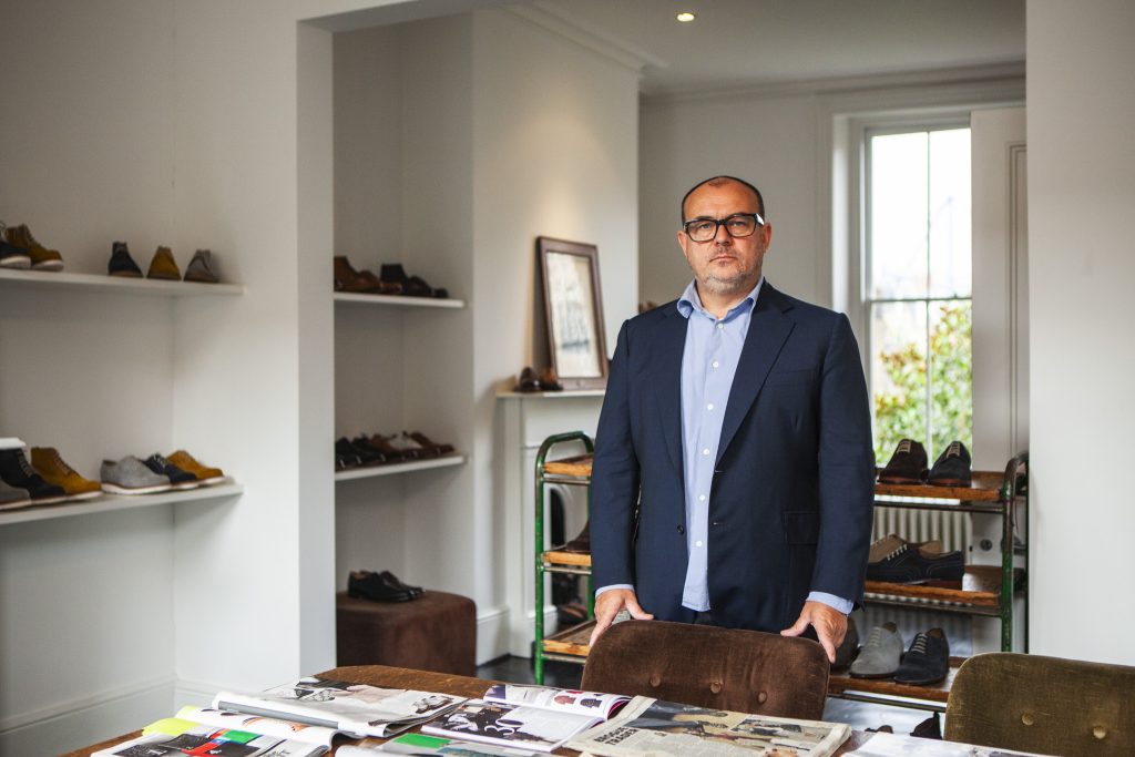 Lifestyle portrait of Tim Little, Creative Director and owner of Grenson and Tim Little with glasses, jacket and shirt standing in a regency style double room in front of a chair and table by portrait photographer Andy Barnham