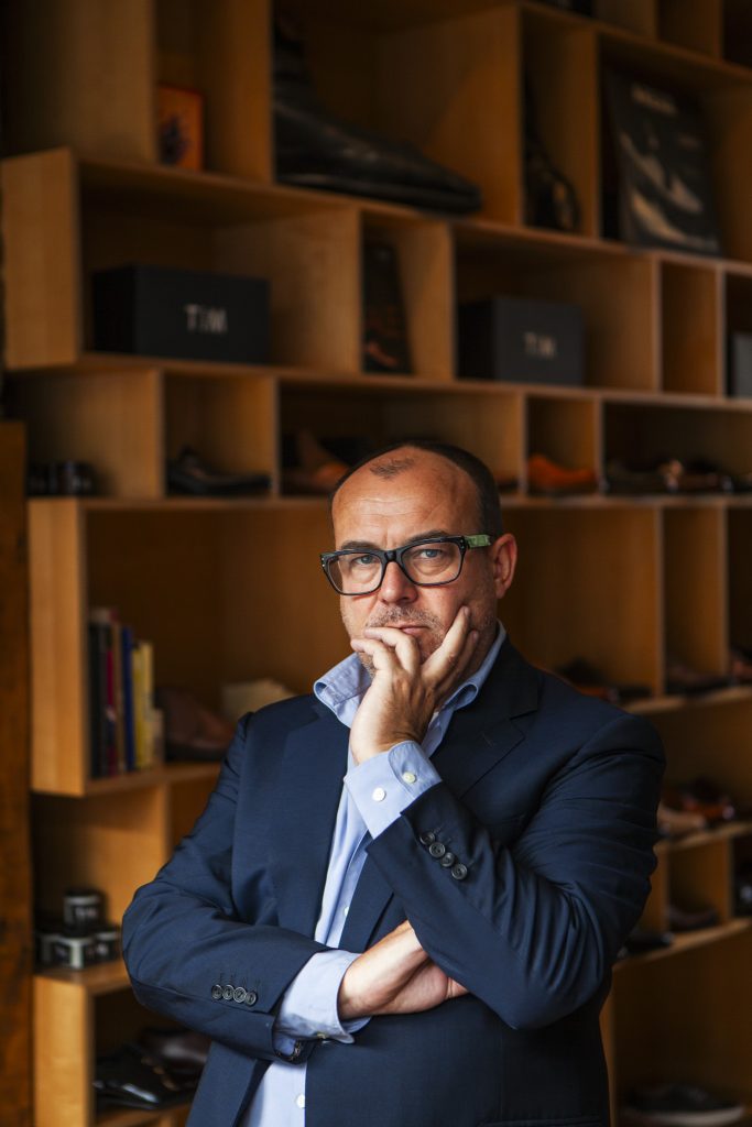 Lifestyle portrait of Tim Little, Creative Director and owner of Grenson and Tim Little with glasses, jacket and shirt standing in front of a shelf of shoes with his chin in his hand looking at the camera by portrait photographer Andy Barnham