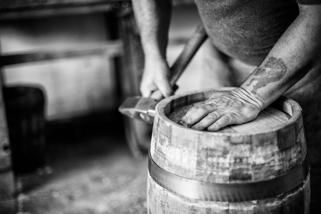 Black and white lifestyle photograph of Master Cooper Alastair Simms, holding a hammer, hand making a wooden cask by portrait photographer Andy Barnham
