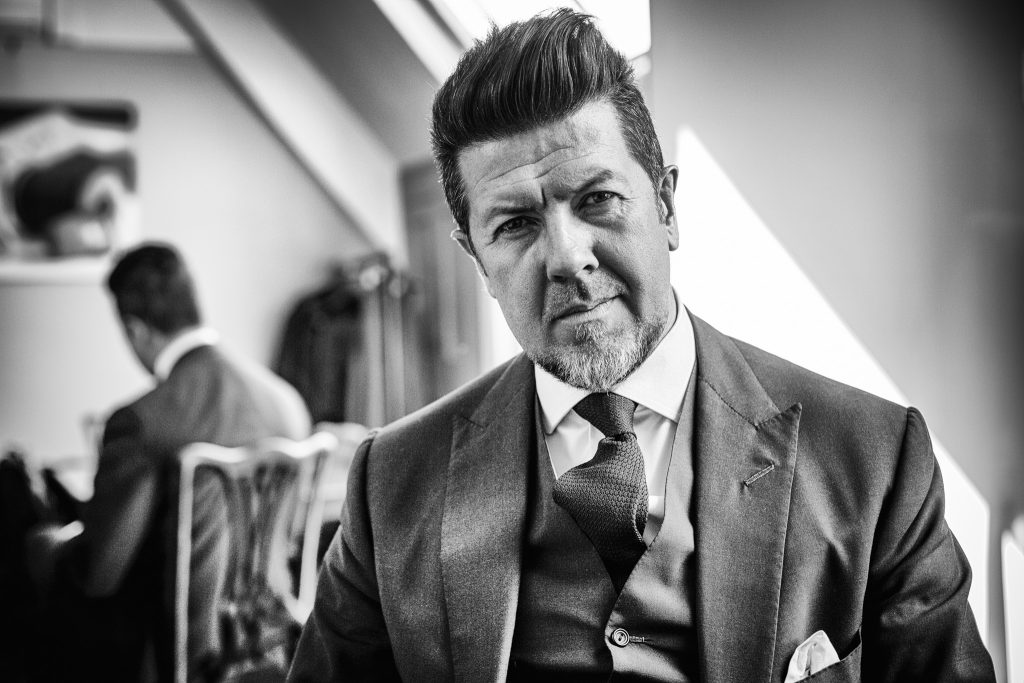 Lifestyle portrait of Savile Row tailor David Ward in three piece suit sitting with reflection of himself in mirror behind by portrait photographer Andy Barnham