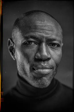 Headshot with analogue border, of Rodney Charles, American & British screenwriter and producer in a black turtleneck jumper in front of a grey background by portrait photographer Andy Barnham