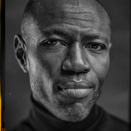 Headshot with analogue border, of Rodney Charles, American & British screenwriter and producer in a black turtleneck jumper in front of a grey background by portrait photographer Andy Barnham