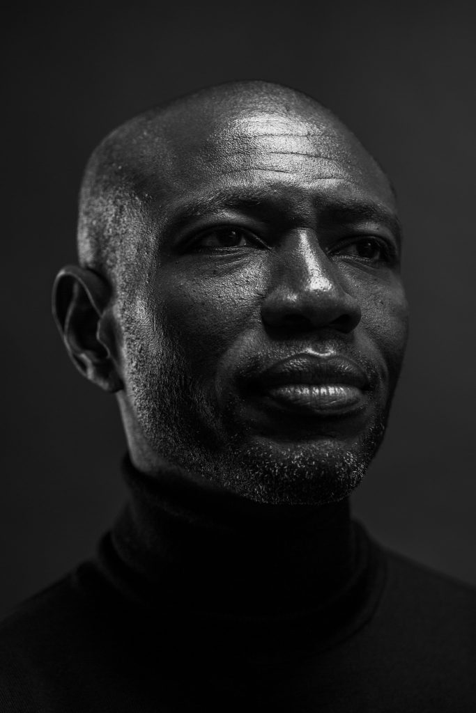 Headshot of Rodney Charles, American & British screenwriter and producer in a black turtleneck jumper in front of a grey background by portrait photographer Andy Barnham