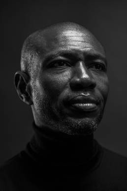 Headshot of an black man in a black turtleneck jumper in front of a grey background by portrait photographer Andy Barnham