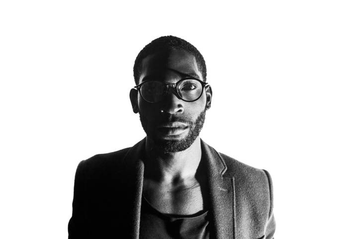 Black and white studio portrait of British rapper and singer Tinie Tempah in glasses and a jacket looking at the camera lit by one light on camera right by portrait photographer Andy Barnham