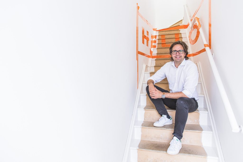 Lifestyle portrait of product designer Christoph Behling in white shirt, jeans and trainers sitting on wooden stairs with hello in orange tape behind him by portrait photographer Andy Barnham