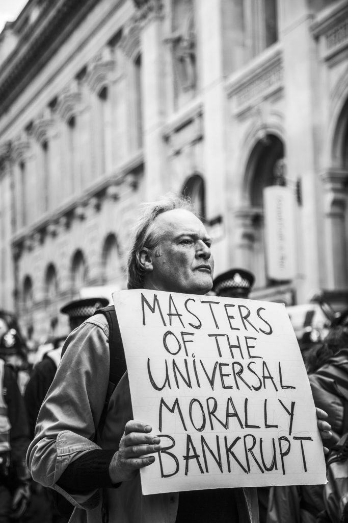 Black and white photo of a man, surrounded by police, holding a sign saying masters of the universal morally bankrupt