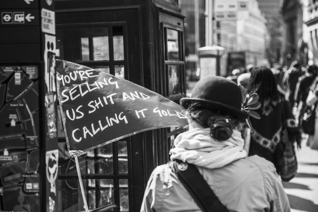 Black and white photo of a woman wearing a bowler hat, next to a phone booth, holding a sign saying you're selling us shit and calling it gold