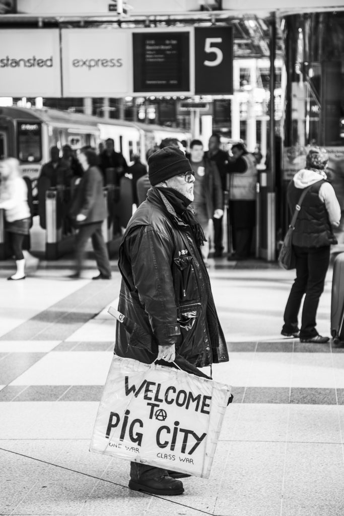 Black and white photo of a man, standing in a train station, holding a sign saying welcome to pig city