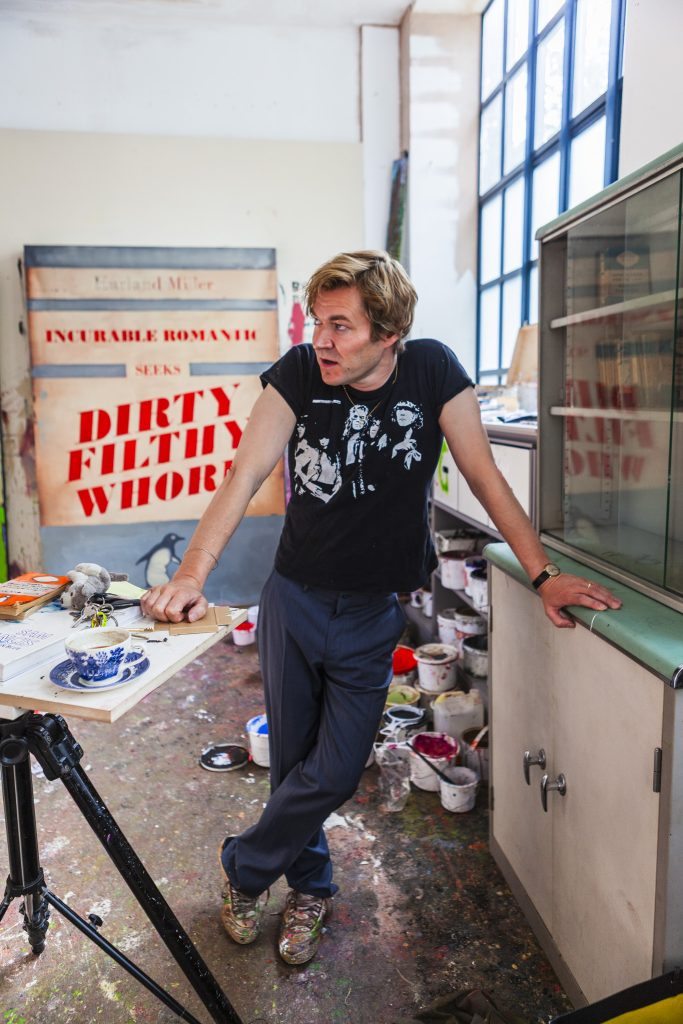 Portrait of British artist Harland Miller in black t- shirt standing in the doorway of an artist studio standing in front of canvas on which Dirty Filthy Whore has been painted by portrait photographer Andy Barnham