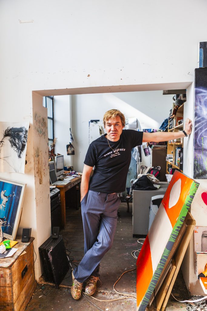 Portrait of British artist Harland Miller in black t- shirt standing in a doorway in his studio surrounded by canvases and painting utensils by portrait photographer Andy Barnham