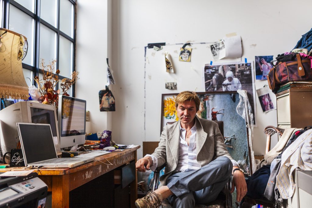 Portrait of British artist Harland Miller sitting in old brown leather chair in his office next to a computer by portrait photographer Andy Barnham