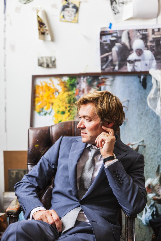 Portrait of British artist Harland Miller in light grey suit sitting in old brown leather chair surrounded in front of a noticeboard filled with reference images  by portrait photographer Andy Barnham