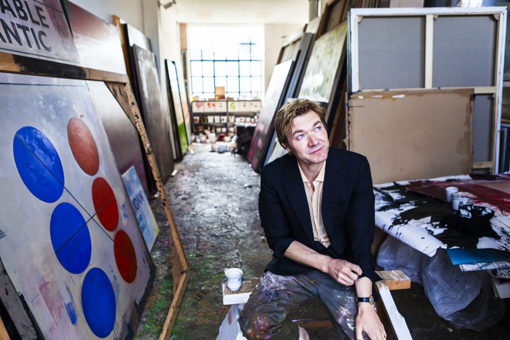 Portrait of British artist Harland Miller sitting on a bench in his painting studio, rolling up his jacket sleeves, surrounded by canvases by portrait photographer Andy Barnham