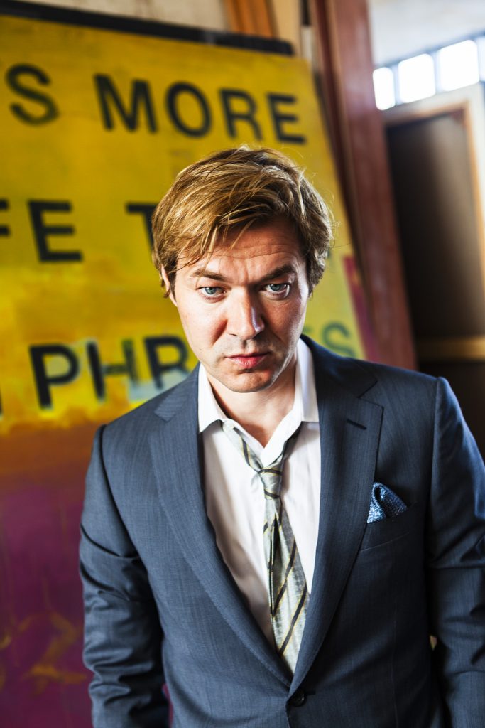 Portrait of British artist Harland Miller in light grey suit standing in front of yellow and fuchsia canvas by portrait photographer Andy Barnham