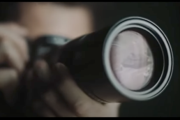 Photographer with a camera lifted to his eye and the reflection of a man standing in front of a tank in the lens