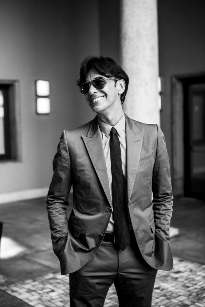 Black and white portrait of former CEO Gucci Patrizio di Marco in suit and sunglasses with his hands in his pockets and smiling, standing in a courtyard by portrait photographer Andy Barnham