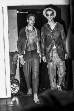 Backstage at Emporio Armani, designed by Giorgio Armani, two male models walk smiling into the backstage area in order to change looks during a fashion show by Andy Barnham