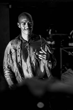 Backstage at Emporio Armani, designed by Giorgio Armani, a male model offers a celebratory hand gesture to the camera the backstage area during a fashion show by Andy Barnham