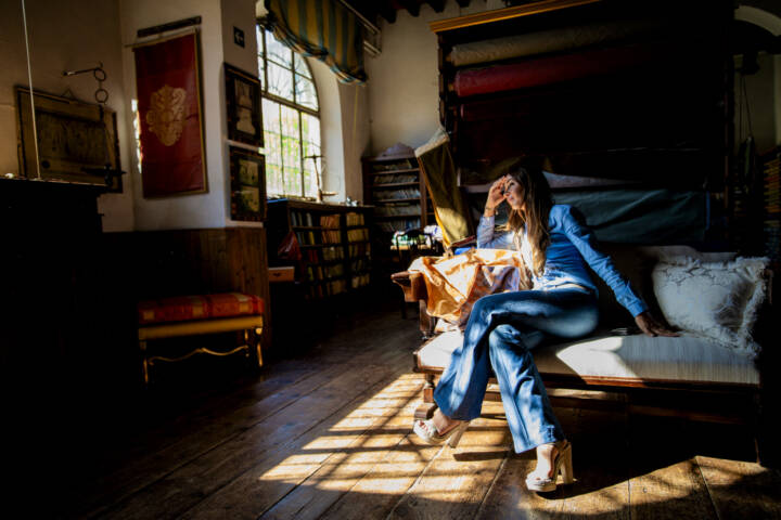 Portrait of woman in blue shirt and jeans sitting on a sofa surrounded by fabrics while sunlight from a window shines on her