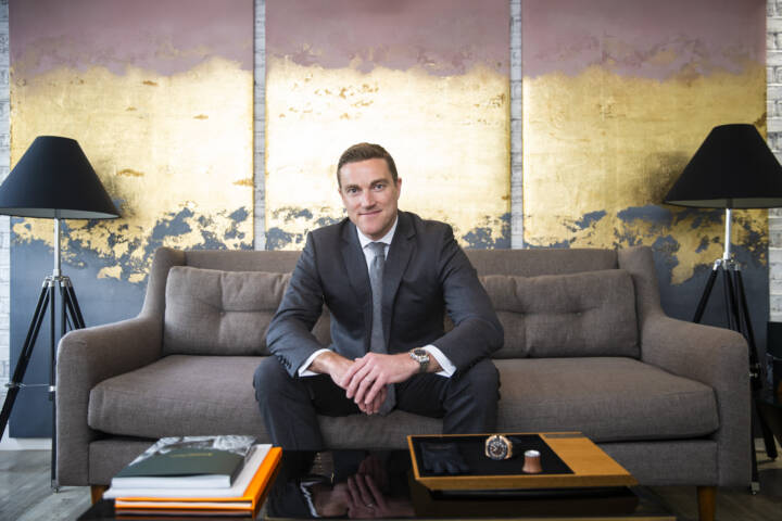 Man in suit sitting on sofa in front of multi coloured triptych work of art with time piece and loupe on table in front of him