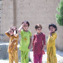Four young girls wearing different colour outfits, three barefoot, standing on a dirt street corner