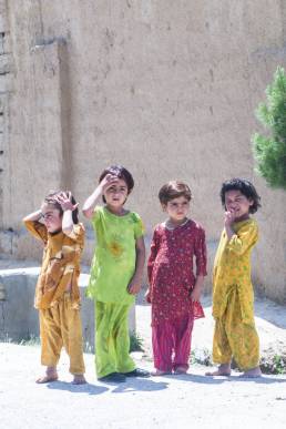 Four young girls wearing different colour outfits, three barefoot, standing on a dirt street corner