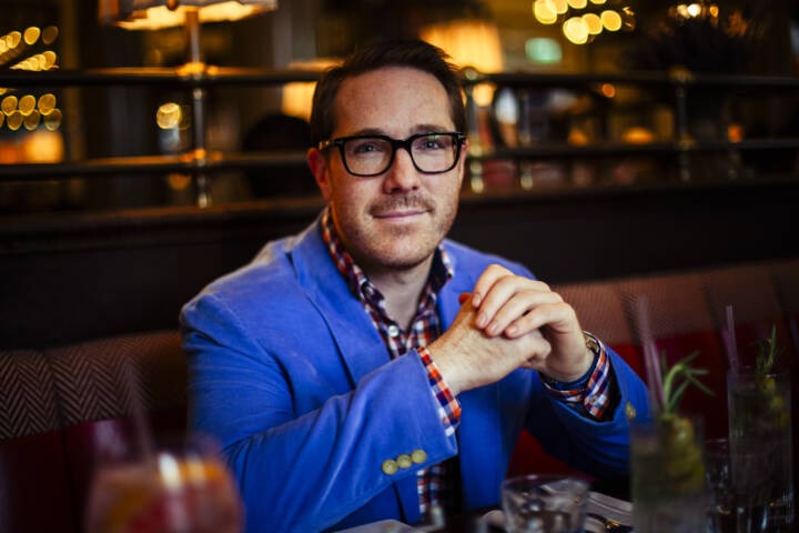 Man in light blue jacket and plaid shirt, wearing glasses with his elbows on a table in a restaurant