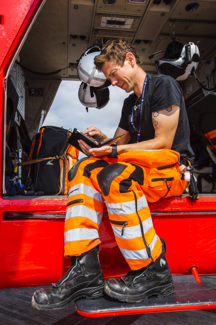 Doctor in high visibility trousers sitting on the side of a helicopter tapping at a small hand held screen.