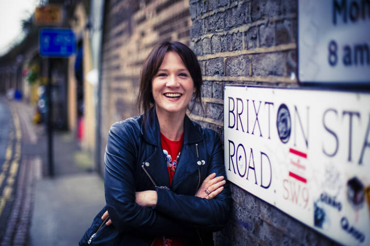 Woman in black leather jacket and red shirt smiling while leaning on a brick wall next to a road sign